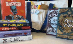 Contents of TeenTober prize pack: a stack of books on the left foreground, a canvas tote bag in the center with buttons and other goodies in and on the pocket, and two books standing up beside the bag.