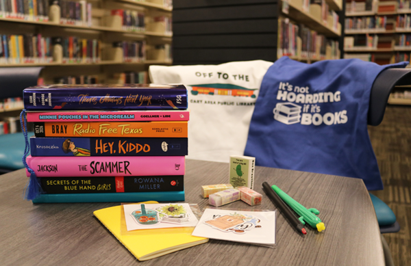 A stack of books with their spines visible sit atop a desk. A small yellow notebooks, sticker packs, three erasers, and two pens surround the book stack. Two canvas bags drape over the chair in the midground. The Teen Area book stacks are out of focus in the background.